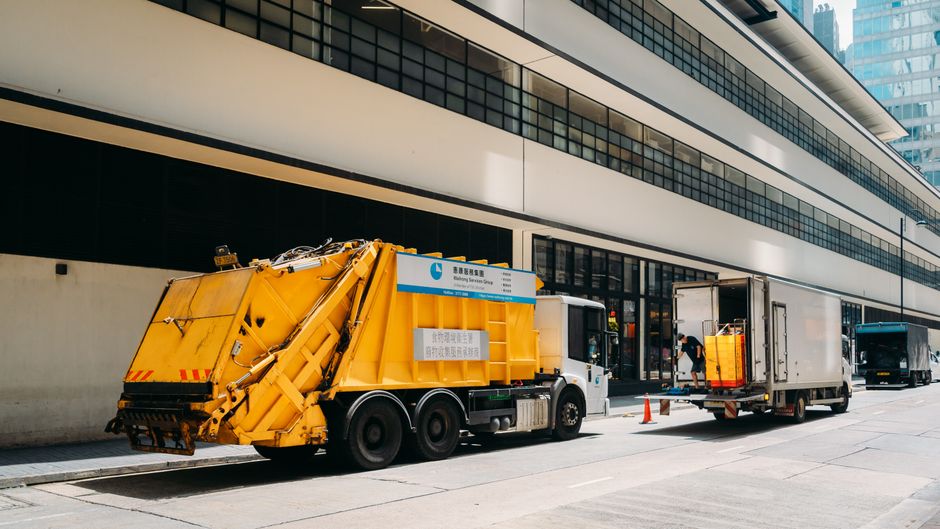 A yellow refuse collection truck with a hydraulic arm is parked on a city street in front of a modern, multi-story office building with large glass windows. The truck is in the process of collecting w