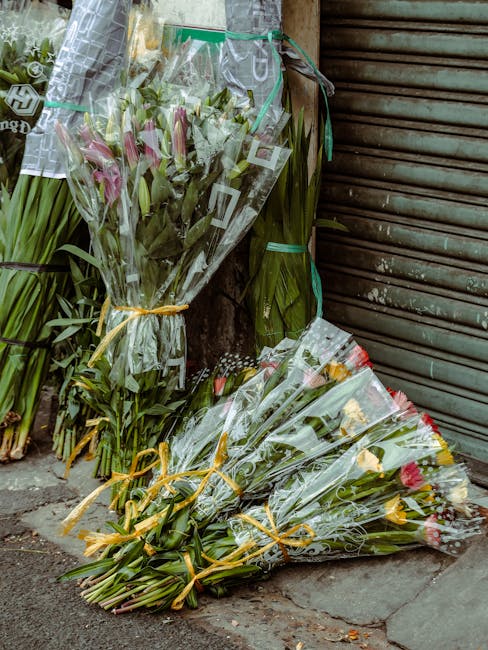 A selection of freshly cut flower bouquets arranged with pink lilies, red and yellow roses, and green foliage, wrapped in clear cellophane and tied with yellow ribbons. The bouquets are resting on the