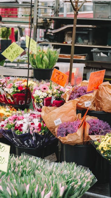 A display of freshly cut flowers at a flower market stall, featuring arrangements of pink, white, and purple tulips in clear plastic wrapping, alongside bouquets of mixed daisies, chrysanthemums, and