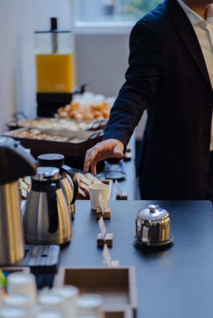 A person in a dark suit and white shirt is reaching for a white paper cup at an office refreshment station, which features a variety of stainless steel coffee pots, a glass juice dispenser filled with