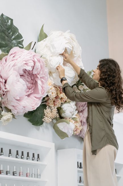 A woman with curly dark hair, dressed in a khaki jacket and beige trousers, is adjusting a large floral wall display in a well-lit indoor setting. The arrangement features oversized peony-like blossom