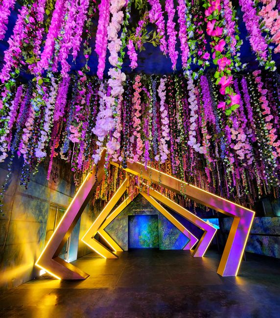 A vibrant floral installation featuring cascading purple and pink flowers hanging from the ceiling, creating a lush canopy. The flowers include wisteria, delphiniums, and cherry blossoms, arranged den