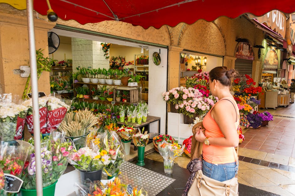A woman with dark hair tied in a bun, wearing an orange sleeveless top and blue jeans, stands in front of a flower stall outside a floral shop. She is holding a small bouquet with both hands, looking 