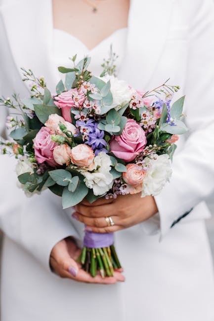 A person dressed in a white outfit is holding a vibrant bouquet of fresh flowers, featuring large pink roses, delicate white blooms, and smaller pastel pink and purple flowers, accented with silvery-g