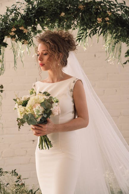 A bride holding a elegant bouquet arrangement featuring white and pale pink peonies, roses, and sprigs of eucalyptus, with a mix of textured petals and lush green foliage. She wears a simple yet sophi